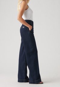 Mujer de pie en jeans de pierna ancha azul oscuro con las manos en los bolsillos, vistiendo una camiseta sin mangas de punto blanca, contra un fondo claro y plano.