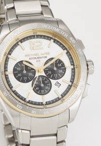 Orologio in acciaio inossidabile con quadrante bianco testurizzato, lunetta con accenti dorati, tre sottocriteri neri e indicazione della data alle 6. Modello: Michael Kors Accelerator.