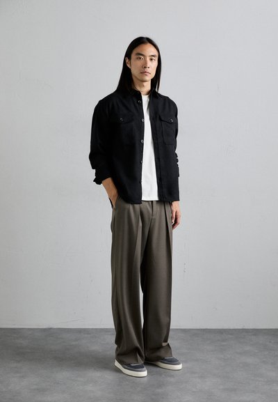 Black button-up shirt over a white t-shirt, paired with gray trousers and gray sneakers. Long hair, neutral background, standing pose.