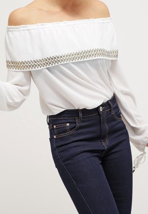 Femme portant un chemisier blanc épaules dénudées avec des clous métalliques sur la poitrine et un jean bleu foncé taille haute.