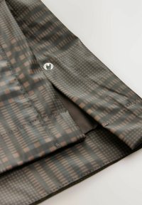 Waterdichte jas gemaakt van een grijs-groene stof met een geometrisch patroon, geaccentueerd door zichtbare stiksels en een metalen drukknoopdetail.