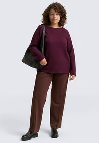 Vínový sveter s dlhými rukávmi a zaobleným výstrihom, doplnený o hnedé nohavice s širokými nohavicami. Modelka nesie čiernu kabelku tote a má na sebe čierne členkové čižmy.