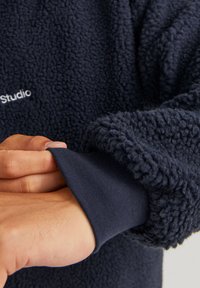 Navy fleece pullover with a textured, fluffy surface. Features a smooth cuff and small white logo on the chest area.
