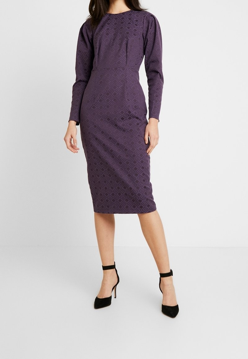 Femme portant une robe violette ajustée, à manches longues, longueur genou, avec un motif subtil en losanges et des escarpins noirs à bride cheville.