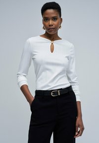 Top blanco de manga larga con textura acanalada, con cuello redondo y detalle de ojal, combinado con pantalones negros y un cinturón negro con hebilla dorada.