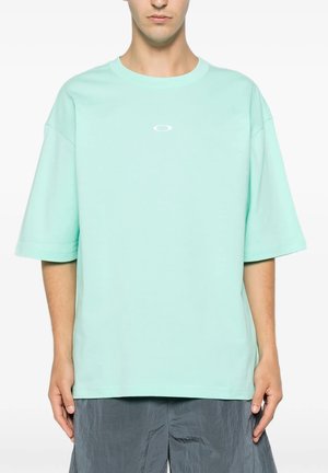 Giovane che indossa una maglietta leggera turchese chiaro a maniche corte, larga, con un piccolo logo bianco sul petto e pantaloni grigi strutturati.