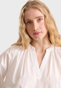 Weiße Baumwollbluse mit V-Ausschnitt und gerafften Schultern. Glatte Textur, lockere Passform und dezente Nahtdetails am Ausschnitt.