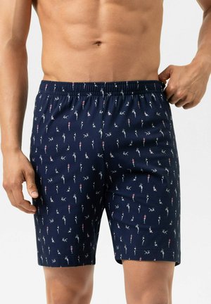 Borstloze man die een navyblauwe short met elastische tailleband aanpast, met kleine witte en rode zwemmerpatronen.