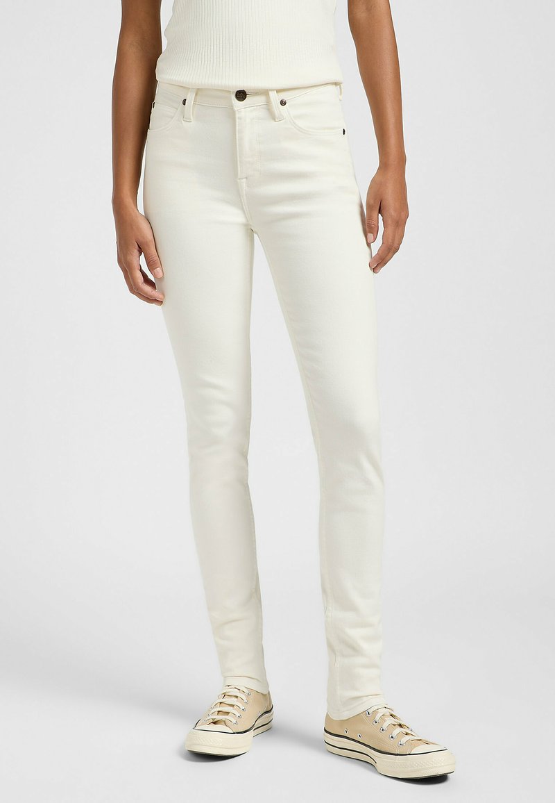 Jeans blancos ajustados de denim, con un clásico diseño de cinco bolsillos, cierre de botón y textura suave. Combinados con zapatillas beige.