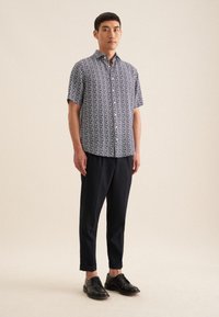 Korte mouwen, overhemd met knopen in een geometrisch blauw en wit patroon, gecombineerd met donkere broek en zwarte schoenen, staande tegen een neutrale achtergrond.