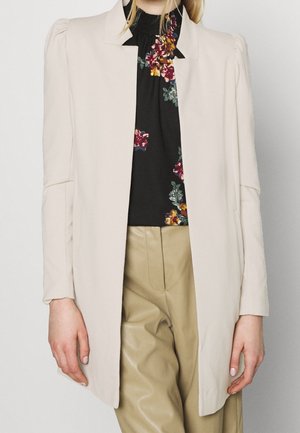 Personne portant un blazer beige sur mesure, un chemisier noir à fleurs rouges, jaunes et vertes, ainsi qu’un pantalon en cuir marron clair, debout devant un fond uni.