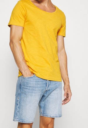 Mann trägt hellgelbes kurzärmeliges Hemd und hellblaue Jeansshorts, mit einer Hand in der Tasche und neutralem Hintergrund.