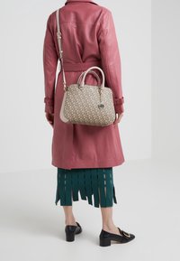 Femme portant un manteau en cuir rose, une jupe verte foncée à franges, des chaussures à talons noires, tenant un sac à main beige monogrammé avec une bandoulière et un porte-clés.