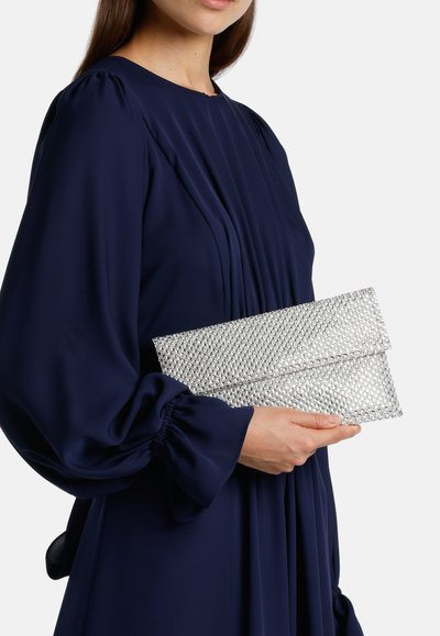 Egy nő tengerészkék ruhát visel, kezében egy csillogó ezüst táskával, amely elegáns ujjakra és finom redőkben gazdag.