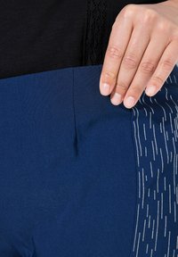 I pantaloni sportivi color navy presentano un tessuto liscio con un pannello laterale testurizzato che mostra un motivo lineare bianco. Una mano tocca l'area della vita.