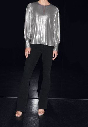 Top lungo metallizzato argento a maniche lunghe con scollatura rotonda e dettaglio a chiave, abbinato a jeans neri a campana e tacchi trasparenti.