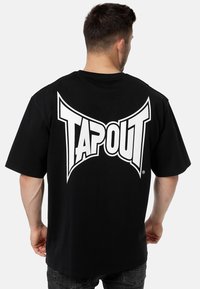 Fekete pamut póló, nagy fehér "TAPOUT" logóval a háton. Rövid ujjú, laza fazonú, sima textúrájú anyag.