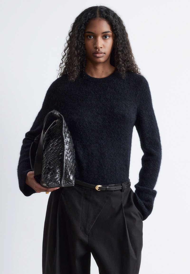 Schwarzer Strickpullover mit langen Ärmeln, kombiniert mit schwarzen hoch taillierten Hosen und einer strukturierten, gewebten schwarzen Handtasche.