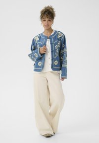 Jeans jakna z vezenimi cvetličnimi vzorci v modrih in kremnih tonih, z gumbnim zapiranjem spredaj in širokimi rokavi, v kombinaciji s bež širokimi hlačami.