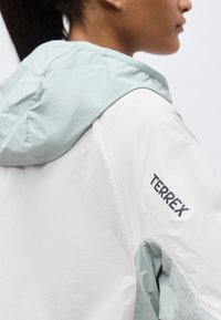 Άτομο που φοράει λευκό και ανοιχτό πράσινο μπουφάν με κουκούλα, με ορατό το λογότυπο «TERREX» στο πάνω μέρος του μανσετιού, στραμμένο πλάγια πάνω σε απλό φόντο.