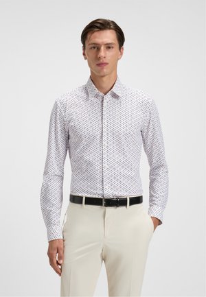 Hombre vestido con una camisa blanca de manga larga con un patrón, metida dentro de pantalones beige con un cinturón negro, posando frente a un fondo blanco liso.