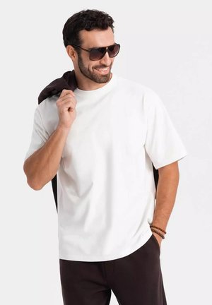 Hvid T-shirt med korte ærmer, afslappet pasform, i bomuldsmateriale. Bruges med brune bukser og et brunt beklædningsstykke over skulderen. Solbriller; lys baggrund.