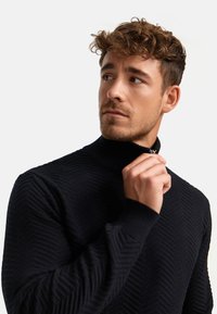 Svart stickad tröja med hög krage och strukturerat fiskbensmönster. Närbild av halvdragkedjedetalj och ribbade ärmslut.