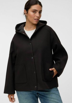Femme portant une veste noire à capuche avec des boutons-pression et des poches avant, associée à un jean bleu, debout devant un fond uni.