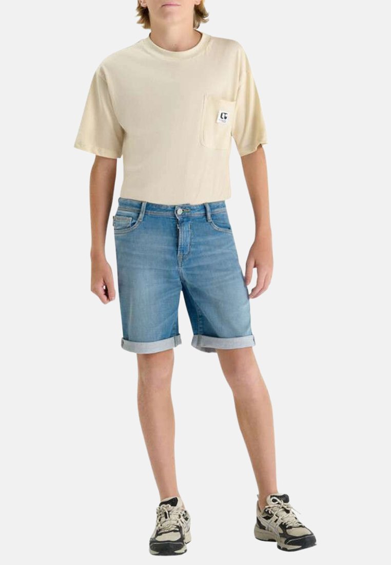 Jeune portant un t-shirt beige à manches courtes avec poche poitrine, un short en jean avec revers roulés et des chaussures de sport beige et noires, debout.