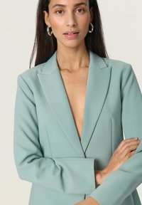 Menta zöld blézer mély V-kivágással, testhezálló fazonban, csúcsos hajtókákkal és hosszú ujjakkal. Sima anyag minimális részletezéssel.