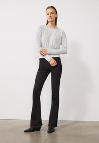 Lichtgrijs geribbeld shirt met lange mouwen, gecombineerd met zwarte uitlopende jeans en zwarte haklaarzen, tegen een effen witte achtergrond.