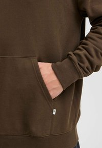 Brauner Sweatshirt mit gerippter Textur, Seitentasche und sichtbaren Nähten. Die Bündchen sind enganliegend und es gibt ein kleines weißes Etikett am Saum.