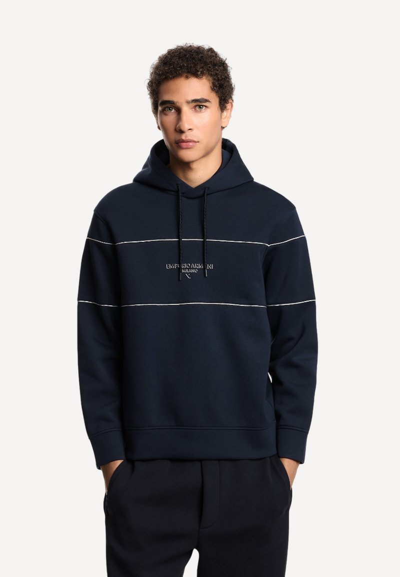 Giovane uomo con capelli ricci che indossa un hoodie blu navy con strisce orizzontali bianche e le mani nelle tasche dei pantaloni, in piedi su uno sfondo semplice.