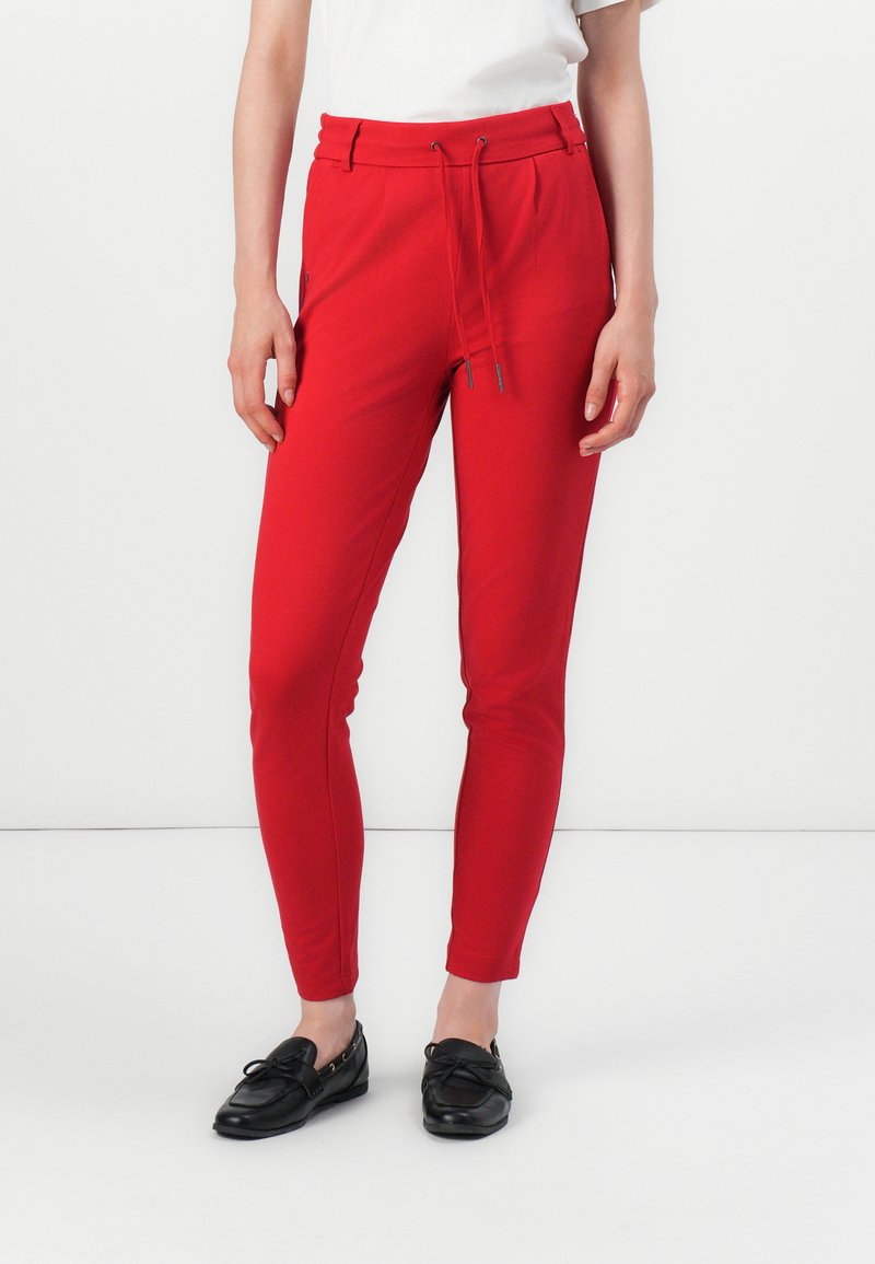 Une femme se tient debout, vêtue de pantalons rouges à cordon de serrage, associés à une chemise blanche et des chaussures noires, le tout sur fond blanc.