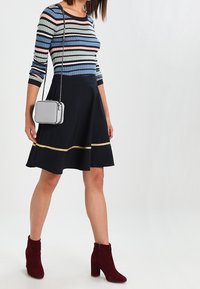 Haut à manches longues rayé en bleu, rose et noir avec un col rond. Jupe noire évasée avec une touche dorée. Une paire de bottines bordeaux.