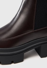 Tmavě hnědá kožená kotníková bota s černou elastickou boční částí, ozdobným prošíváním a robustní černou gumovou podrážkou s hlubokými drážkami.