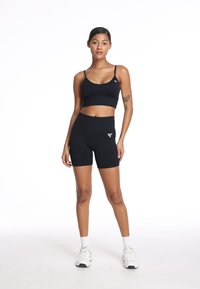 Czarny stanik sportowy i wysokie szorty, wykonane z elastycznego materiału. Szorty mają logo. Model nosi białe buty sportowe.