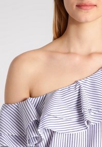 Top a spalla scoperta a righe con design a balze in bianco e blu navy. Presenta un tessuto texturizzato con vestibilità morbida e scollatura delicata.