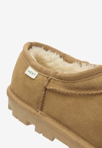 Tan suede hjemmesko med en blød cremefarvet foring, rund tå og tekstureret gummisål. Har et lille mærke med "NEXT" på siden.