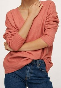 Różowy koralowy sweter z dzianiny z dekoltem w serek, z prążkowanymi mankietami i luźnym krojem, w zestawieniu z niebieskimi dżinsami z wysokim stanem, prezentującymi miękką teksturę.