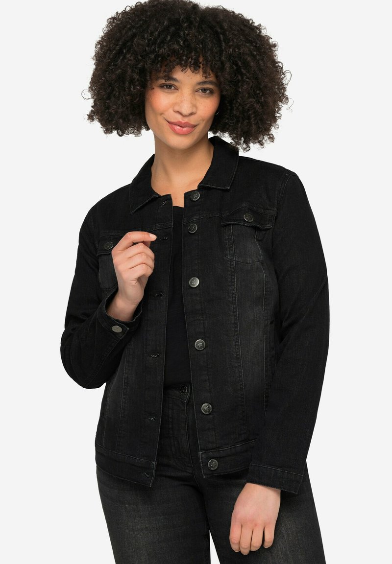 Veste en denim noir à manches longues, fermée par des boutons, avec deux poches poitrine et une texture légèrement usée. Portée par-dessus un haut noir.