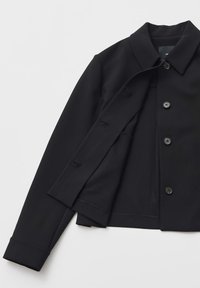 Zwarte jas van gladde stof, voorzien van een klassieke kraag, knopen aan de voorkant en lange mouwen met knoopmanchetten. Gevoerde binnenkant.