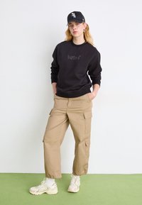 Czarna bluza z logo, beżowe bojówki z kieszeniami, białe skarpetki i beżowe sneakersy. Noszona z czapką w kolorze czarnym. Stoi na zielonej powierzchni.