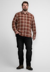 Man staat tegen een witte achtergrond, gekleed in een bruine geruite overhemd, zwarte jeans en bruine veterschoenen, met één hand in zijn zak.