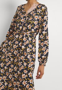 Květinové šaty s námořnickým pozadím a růžovými a žlutými květinami, V-výstřihem, dlouhými nadýchanými rukávy a shrnutým pasem.