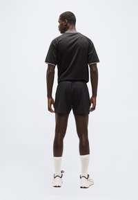 Άνδρας φοράει μαύρη αθλητική εμφάνιση, λευκές κάλτσες και λευκά αθλητικά παπούτσια, στέκεται με την πλάτη γυρισμένη προς τα εμπρός σε απλό ανοιχτό φόντο.