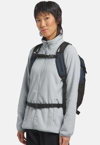 Giacca in fleece azzurro chiaro con colletto alto, zip frontale e orlo elasticizzato. Sono visibili anche delle cinghie nere regolabili e uno zaino blu scuro.