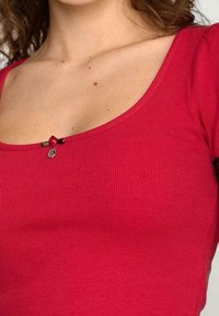 Rød ribbet top med en scoop-halsudskæring, dekoreret med en lille knapaccent i midten. Stoffet er blødt og tætsiddende.