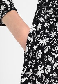 Tissu noir avec un motif floral blanc, révélant une main dans la poche d'un vêtement à manches courtes et à coupe décontractée.