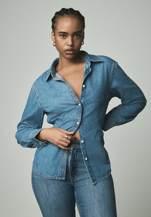 Denimhemd mit durchgehender Knopfleiste, spitzen Kragen und langen Ärmeln. Hellblaue Farbe mit weicher Textur. Teilweise geöffnet getragen.